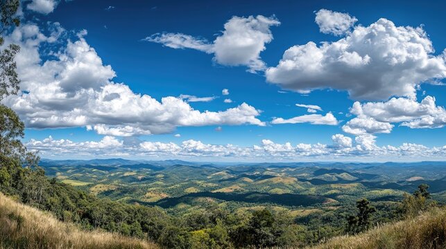 Vibrant panoramic landscape featuring a bright blue sky dotted with fluffy white clouds over lush green hills and rolling mountains.