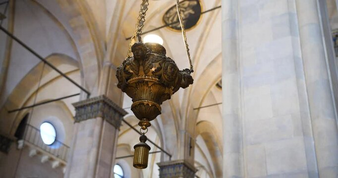 An ornate hanging lantern in a cathedral interior, featuring detailed carvings and suspended by chains, with arched ceilings and columns in the background.