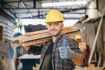 Smiling construction worker in a yellow hardhat and plaid shirt holding wood planks on his shoulder...