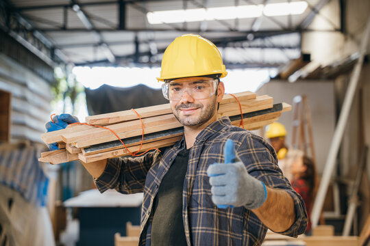 Smiling construction worker in a yellow hardhat and plaid shirt holding wood planks on his shoulder and thumb up finger. Ideal for carpentry, woodworking in furniture workshop, National Carpenters Day - Powered by Adobe