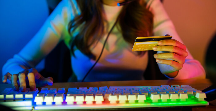 Asian gamer playing online video game and holding a credit card for payment game online, young woman in gaming headphones using computer for playing games at home, broadcast streaming live, Front view