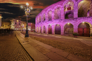 The Arena di Verona, illuminated in fuchsia hues against a vibrant pink sunset sky. Long exposure evening shot capturing the motion blur of people passing by.