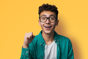 Excited surprise shock, happy young man wear braces, tooth brackets, eye glasses, open mouth, say yes hand gesture, isolated yellow background. Sale, win lottery, dental care, ophthalmology ad concept