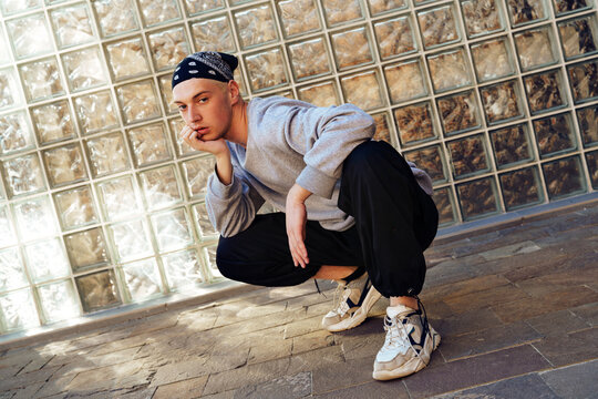 A young man wearing a gray sweatshirt and black pants squats in front of a glass block wall