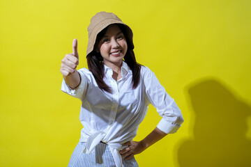 An Asian woman wearing a beige hat, white shirt, and striped pants smiles brightly against a yellow...