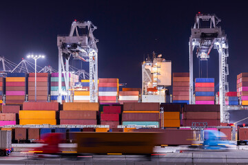 Cranes unload intermodal containers from a ship at port, with motion blurred semi trucks rush in...