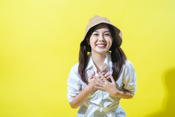 An Asian woman wearing a beige hat and a white shirt with striped pants stands against a yellow background, looking surprised with her hands on her chest and her mouth open in amazement.