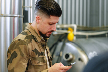 A man in a camo shirt uses his phone while standing in a factory