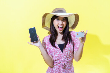 Asian woman in a pink floral dress, wearing a wide-brimmed straw hat, holding a smartphone in one hand and a credit card in the other, smiling brightly against a vibrant yellow background