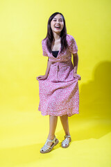 Asian woman in a pink floral dress holding its hem playfully, smiling warmly and posing confidently against a vibrant yellow background, showcasing a cheerful and stylish summer look.