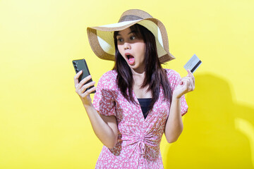 Asian woman in a pink floral dress, wearing a wide-brimmed straw hat, holding a smartphone and a credit card with a surprised expression against a vibrant yellow background, showcasing shock