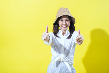 An Asian woman wearing a beige hat, white shirt, and striped pants smiles brightly against a yellow background, giving two enthusiastic thumbs up as a gesture of positivity and approval.