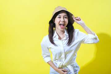 An Asian woman in a white shirt and striped pants poses cheerfully against a yellow background. She wears a beige hat, smiles widely, and has her hand on her hip, exuding vibrant energy.