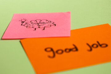 Close up of pink and orange sticky note paper with drawing of brain holding a light bulb and written message saying good job on a mint green background