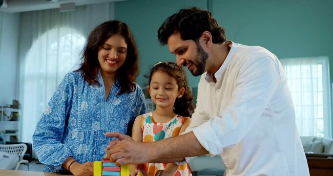Indian Asian young married couple enjoying quality time with their adorable daughter, building a home using colorful wooden block toys at a table in their modern home