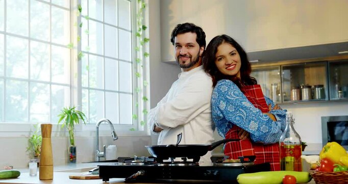 Indian Asian couple in a modern kitchen, young wife and husband enjoying cooking together, posing back-to-back with thumbs up and smiles for the camera