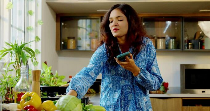 Young beautiful Indian Asian woman cooking food in a pan in the kitchen while speaking on a speakerphone, multitasking and managing kitchen tasks efficiently