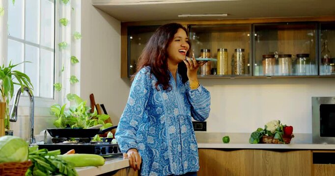 Young beautiful Indian Asian woman cooking food in a pan in the kitchen while speaking on a speakerphone, multitasking and managing kitchen tasks efficiently
