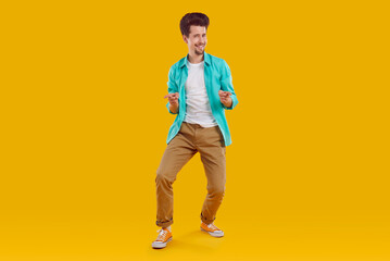 Fototapeta premium Hey you, yes you. You're going to the party. Full body length happy handsome young man in casual turquoise shirt and brown pants standing on yellow background, pointing fingers at you and smiling
