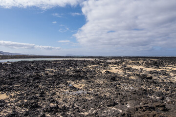 White sand beach with solidified volcanic lava flows, black rocks. Mountains in the background and sky with big white clouds. Caletón Blanco, Lanzarote, Canary Islands, Spain