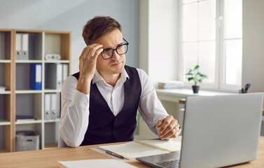 Confident young attractive business man in suit and glasses looking seriously at computer monitor...