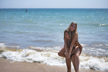 Two friends, young and beautiful, one brunette and the other blonde, wearing bikinis, piggybacking each other in a fun and playful attitude, on the beach. Concept beauty, beach, sun, sand, summer.