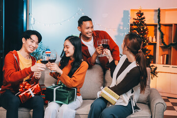 Group of young Asian man and women as friends having fun at a New Year's celebration, holding gift boxes standing by Christmas tree decoration, midnight countdown Party at home with holiday season.
