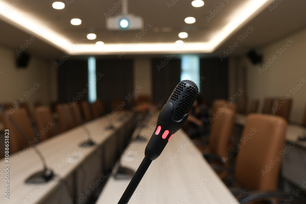 Poster microphone stands ready in empty conference room, with rows of chairs and projector in background, c