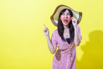 A cheerful woman in a pink floral dress, wearing a wide-brimmed hat, playfully holds pink sunglasses to her mouth while pointing upward, set against a bright yellow background.