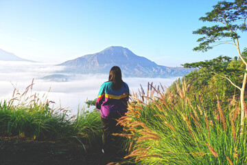 Young woman standing alone among the grass flowers from behind against the mountain view of Mount Batur with blue sky and the fog background. Desa Pinggan - Kintamani, Bali, Indonesia