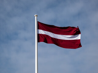 The national flag of Latvia in the wind with cloudy sky. Latvian flag - carmine red with white horizontal stripe