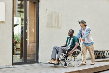 Full length shot of African American man using wheelchair going into cafe in city setting with friend assisting copy space