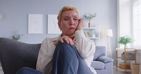 Portrait of a serious and sad elderly woman seated on a couch, lost in deep sadness and thought. Her expression reflects grief and depression, capturing a moment of emotional struggle at home.