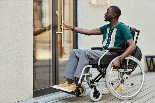 Side view full length portrait of adult African American man with disability using wheelchair and opening door in city copy space