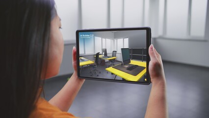 Asian female designer uses tablet computer and virtual reality interior design app to design office workspace. Tablet display shows AI builder software scanning and creating design with 3D furniture.