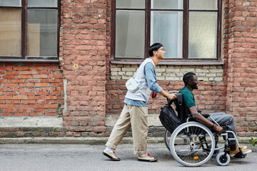 Full length side view of young Asian man assisting friend with disability and pushing wheelchair against brick wall in city copy space