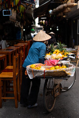 The street vendor with bike loaded of tropical fruits in old town street in Hanoi, old houses and street activites on background