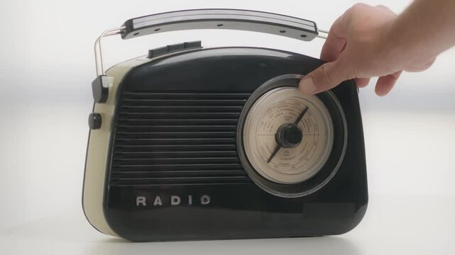 A hand adjusts the dial on a classic vintage black radio, capturing nostalgia and retro design. The radio showcases mid-century style and simplicity, evoking memories of old broadcasts and timeless mu