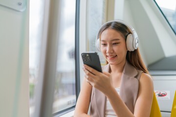 Asian Woman Using Phone and Headphones Listening To Music and Playing Games While Commuting To Work in a Public Train