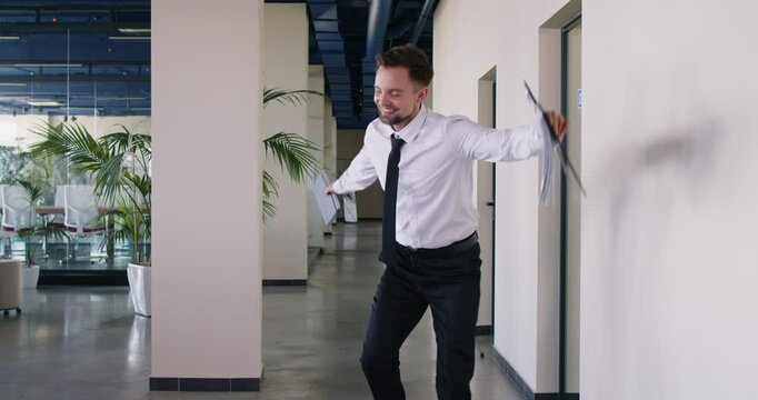 Happy company employee joyfully spinning with clipboard and notebook in his hands and energetically dancing. Jubilant businessman celebrating making successful business deal or achievement in work.
