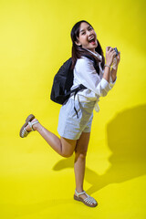 Asian woman with pigtails wearing a white shirt, and a black backpack, holding a camera and smiling enthusiastically, exuding energy and playfulness against a bright yellow background