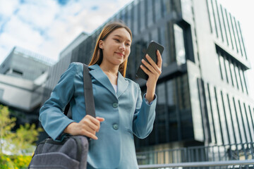 Fototapeta premium Asian Businesswoman Carrying Laptop Bag Using Phone In Front Of A Modern Office Building