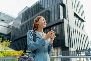 Fototapeta premium Asian Businesswoman Carrying Laptop Bag Using Phone In Front Of A Modern Office Building