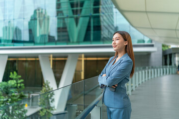 Young Businesswoman Holding Phone and Standing In Front A Modern Office Building, Looking Contented