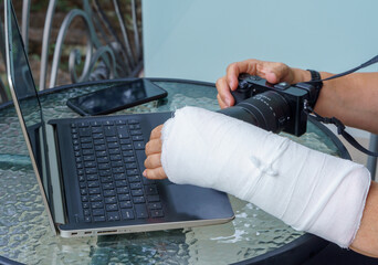 Photographer with broken arm in white plaster cast holding big black camera. Freelancer with broken wrist typing on laptop computer on keyboard. There is a mobile phone on the glass table.