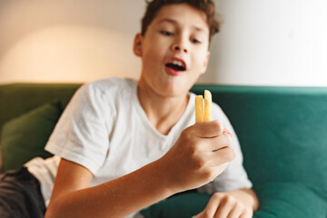 Close up portrait of a satisfied cute boy eating french fries at home on sofa while reading or doing homework. Unhealthy food concept, close up