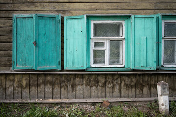 Windows in an old wooden house. Open and closed shutters on the windows. Weathered, faded paint on the wall of the house and on the shutters. Wooden houses in small provincial towns and rural areas.