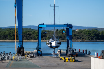 A boat hoist crane holds a yacht for maintenance at a marina. The blue gantry crane, designed for...