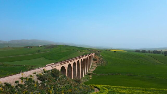 The scene is an aerial view of a bridge crossing over a valley with a train track running through it. On the other side of the valley, there are green fields and mountains in the distance.