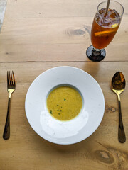 Top view of a white round plate with yellow soup and fork and spoon on it's sides on top of a brown wooden table